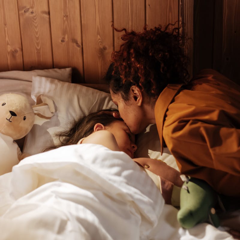 A mother lovingly kisses her sleeping daughter goodnight, creating a tender moment in a cozy, wooden bedroom.